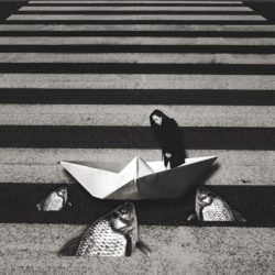 A surrealistic image: on a crosswalk, there is a paper boat, fish coming our of the tarmac and a lady looking down.