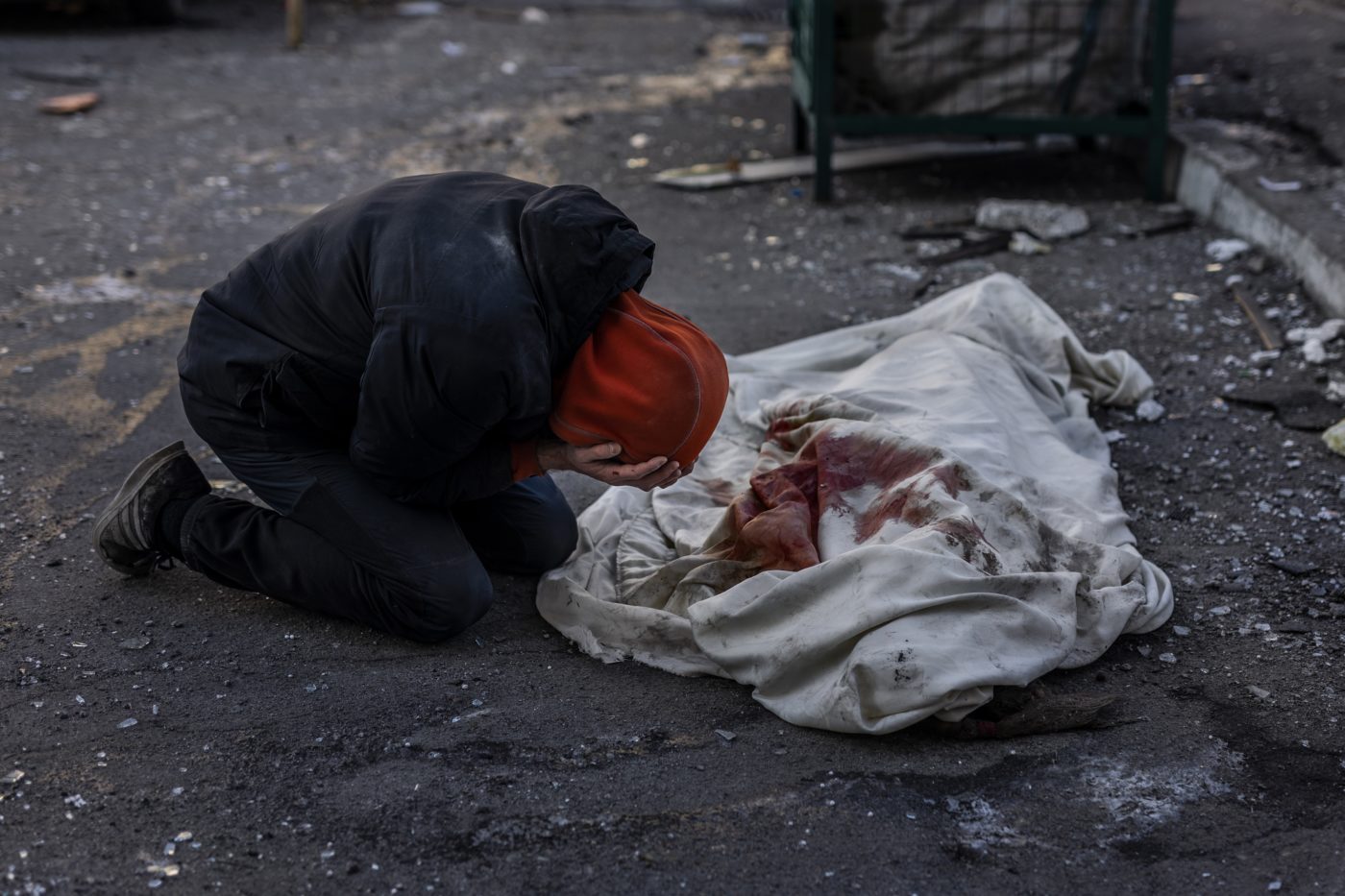 A person on their knees is hunched over the covered and bloody body of a loved one.
