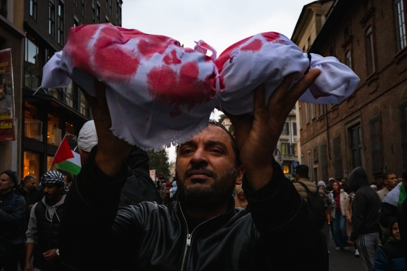 A man surrounded by a group holding Palestinian flags holds up a bloodied sheet.