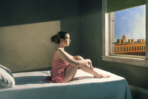 A woman in a pink dress sits in a drab room staring out the open sunny window.